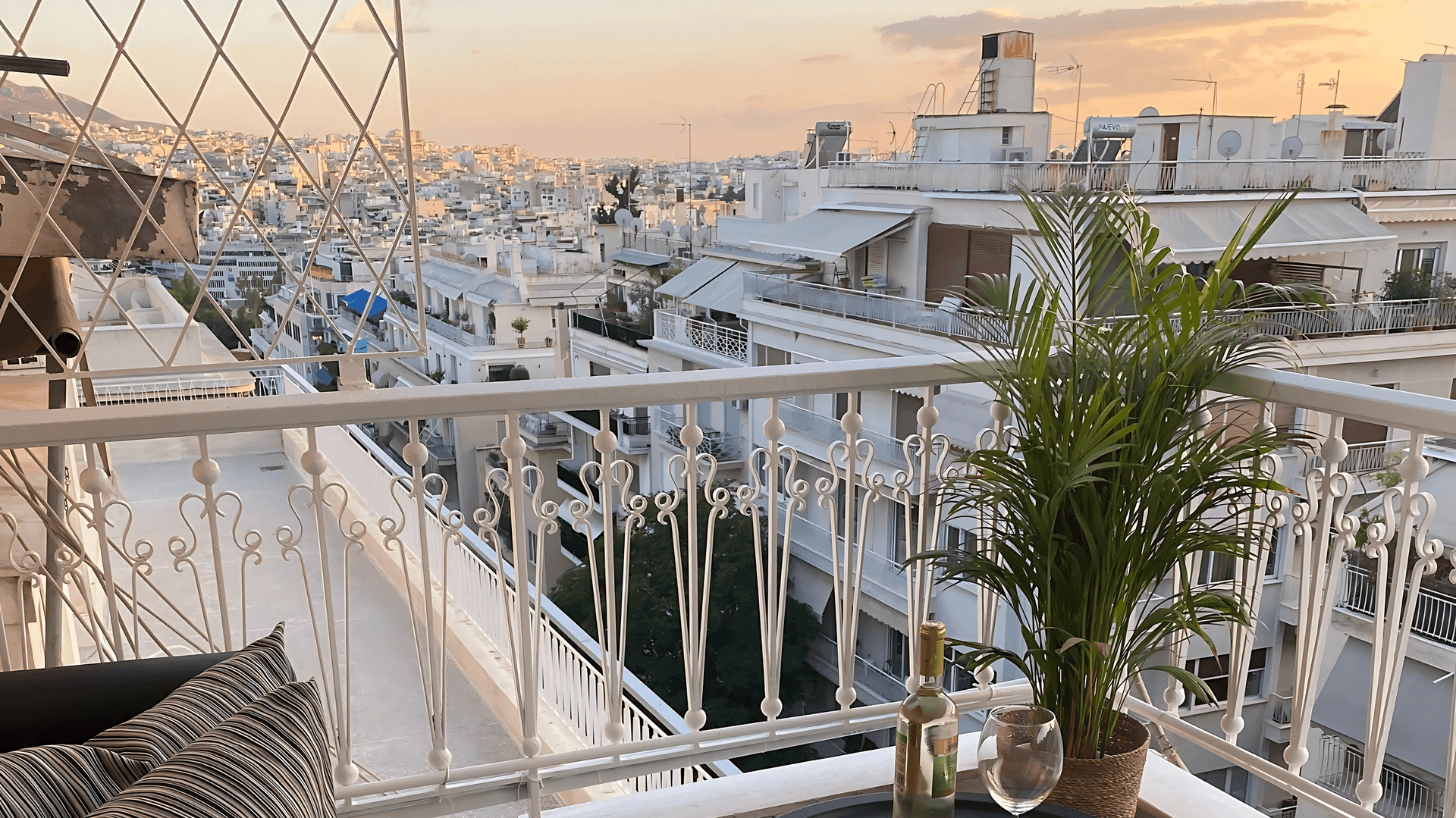 Balcony with potted plant and wine bottle overlooking a cityscape of white buildings at sunset.